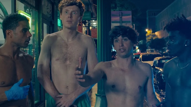 Four shirtless young men stand on a neon-lit Miami street at night, looking startled
