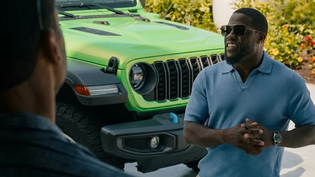 Kevin Hart, in a blue polo and sunglasses, smiles beside a bright green Jeep while talking to a younger man