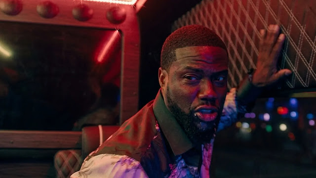 Kevin Hart, one hand raised, reacts inside a neon-lit party bus at night