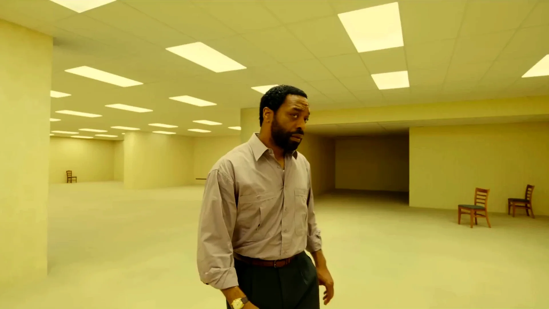 Clark (Chiwetel Ejiofor) stands alone in a vast empty room lit by yellow fluorescent panels, with a few chairs on the carpet