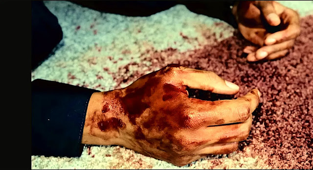 A blood-covered hand in pale gravel at a crime scene in Blood Sacrifice