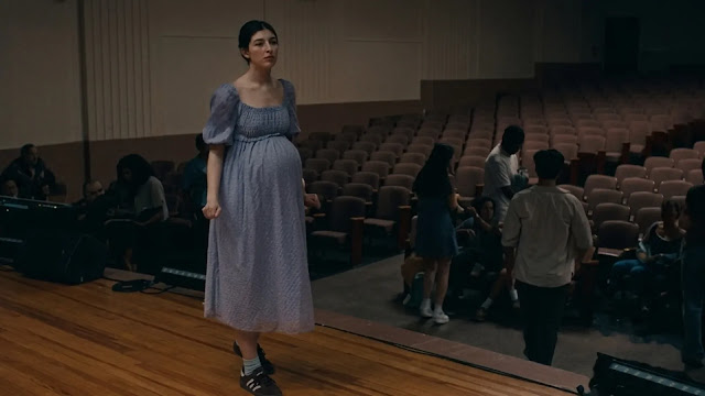 Sophie Birenbaum (Sunny Sandler) standing alone on a school auditorium stage in a pale period dress in Don't Say Good Luck