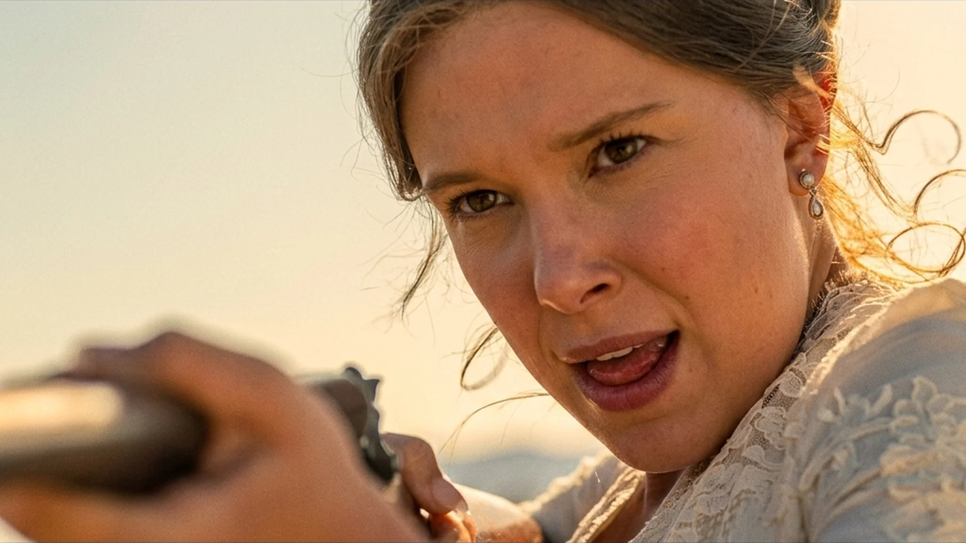 Millie Bobby Brown as Enola Holmes in a wedding dress, flinching as a gun is leveled at her