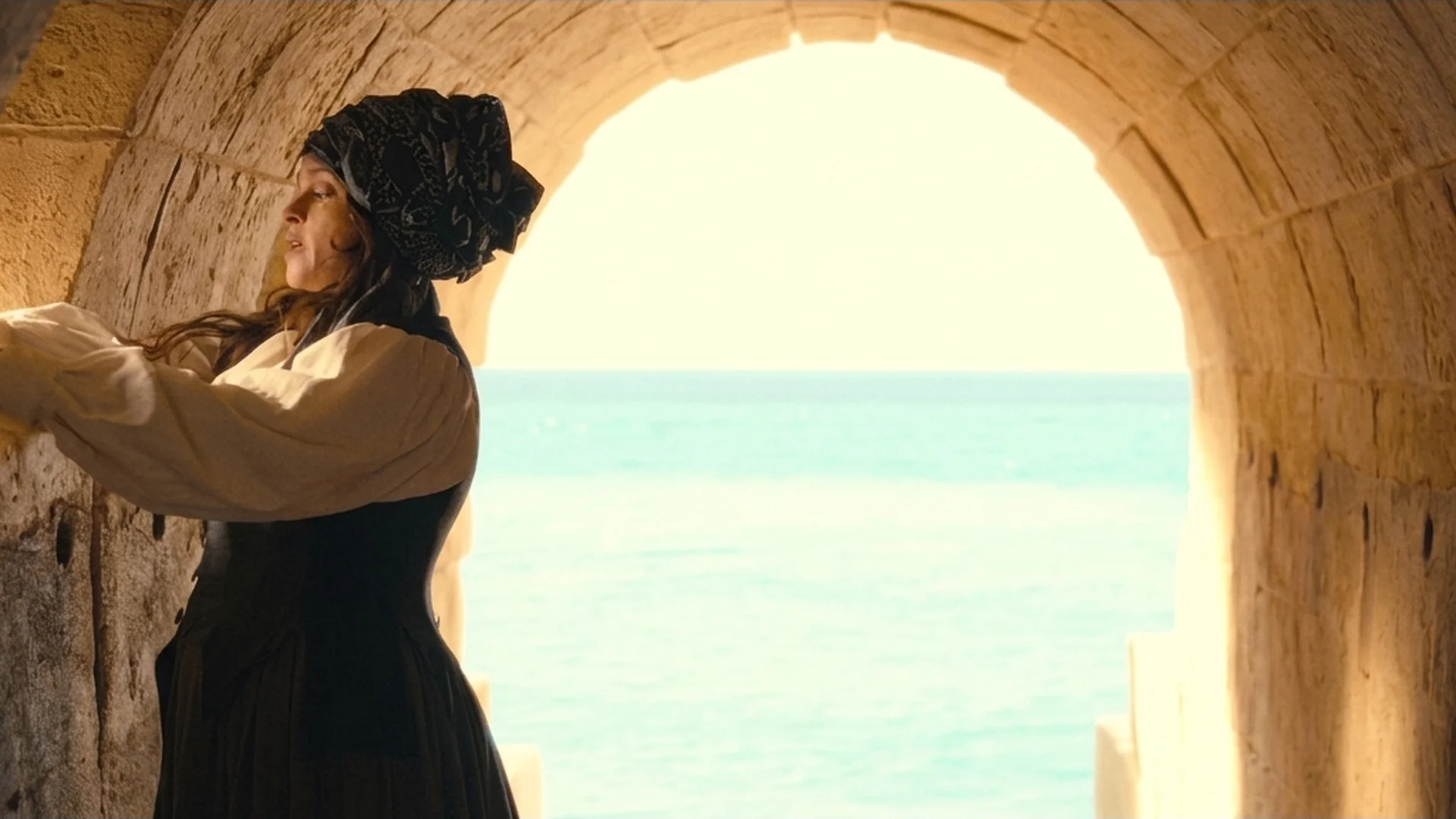 A woman in a headscarf in a sunlit stone archway in Malta, the sea behind her