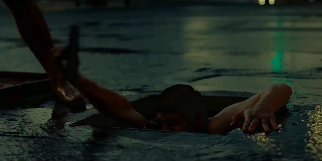 : A man pulls himself up out of an open manhole onto a wet street at dawn