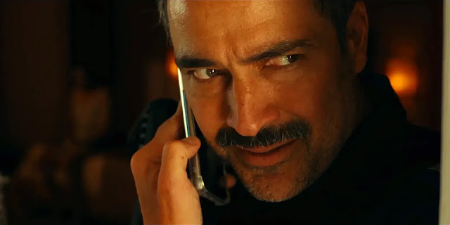 A mustached federal officer talks on a phone in a warm, dim room