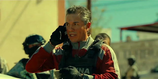 A federal officer in a red jacket talks on a phone beside armed police on a street