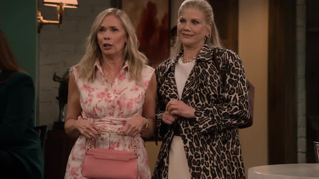 Two women standing side by side in a living room in Leanne, one in a floral dress holding a pink handbag and the other in a leopard print coat