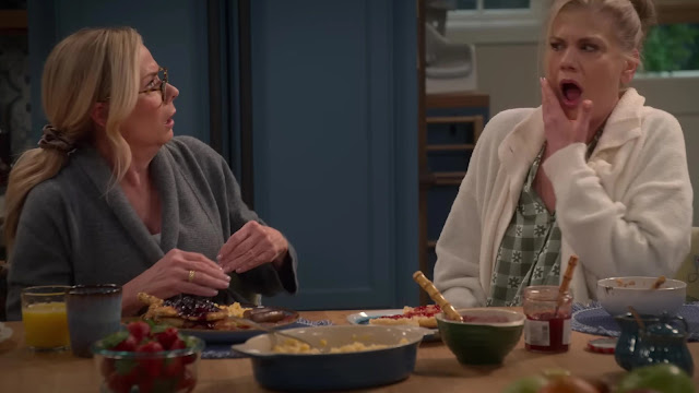 Two women talking across a breakfast table in Leanne, one in glasses and a grey robe, the other reacting with her hand raised