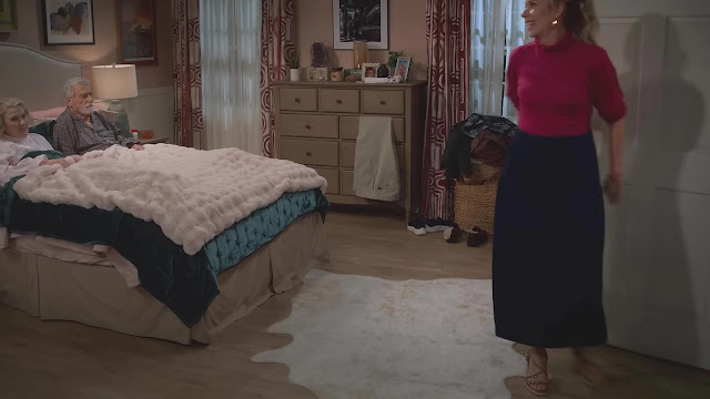 An older couple in bed while a woman in a red top crosses the bedroom in Leanne