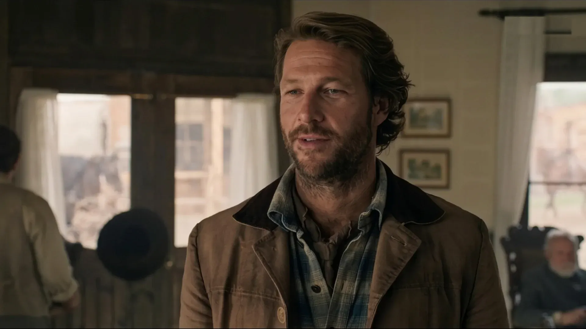 A bearded man in a brown jacket stands indoors in Little House on the Prairie.