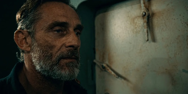 A grey bearded man looking off to one side beside a weathered steel door in Mutiny, rust spreading around its latches