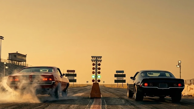 Two muscle cars idle side by side at a nighttime drag race, a starting light between them.