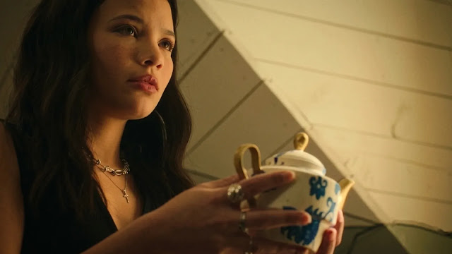 Jackie holds a teacup indoors, looking away in thought.