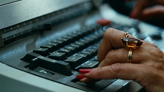 A hand with red nails and a gold ring typing on an office typewriter in 9 to 5