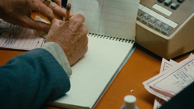 A hand in a suit cuff writing in a notepad beside an adding machine and a stack of invoices in 9 to 5
