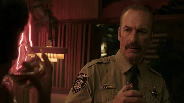 Sheriff Ulysses (Bob Odenkirk) in uniform inside a bar lit by red neon in Normal