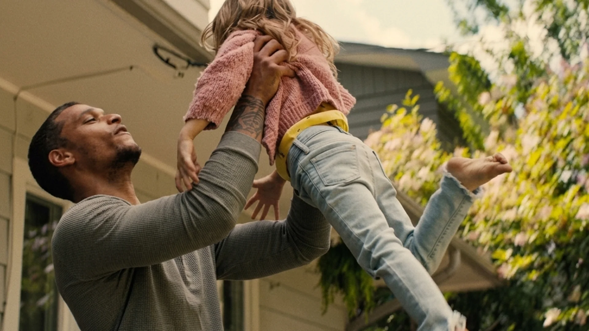 Ledger lifts a laughing little girl overhead in a sunlit backyard in Reminders of Him