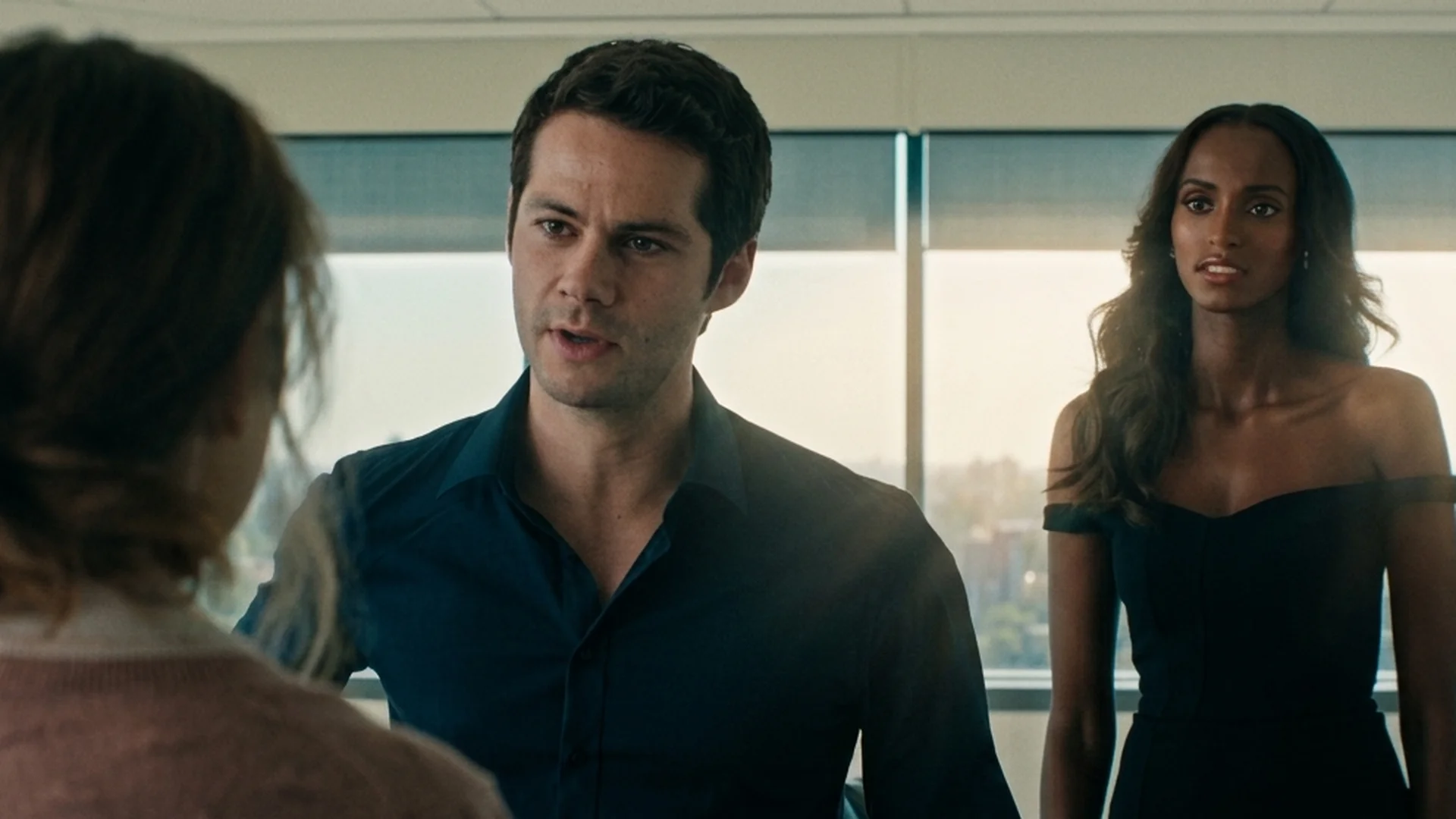 Dylan O'Brien as Bradley stands talking in a glass-walled high-rise office, a woman in a black off-the-shoulder dress beside him, the back of Linda's head in the foreground in Send Help