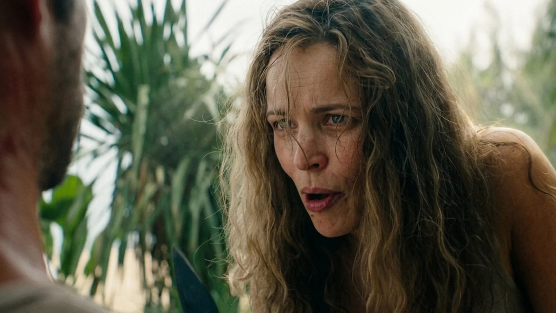 Close-up of Linda with wind-blown hair and an alarmed, open-mouthed expression, a man's shoulder facing her in the foreground, palm fronds behind in Send Help