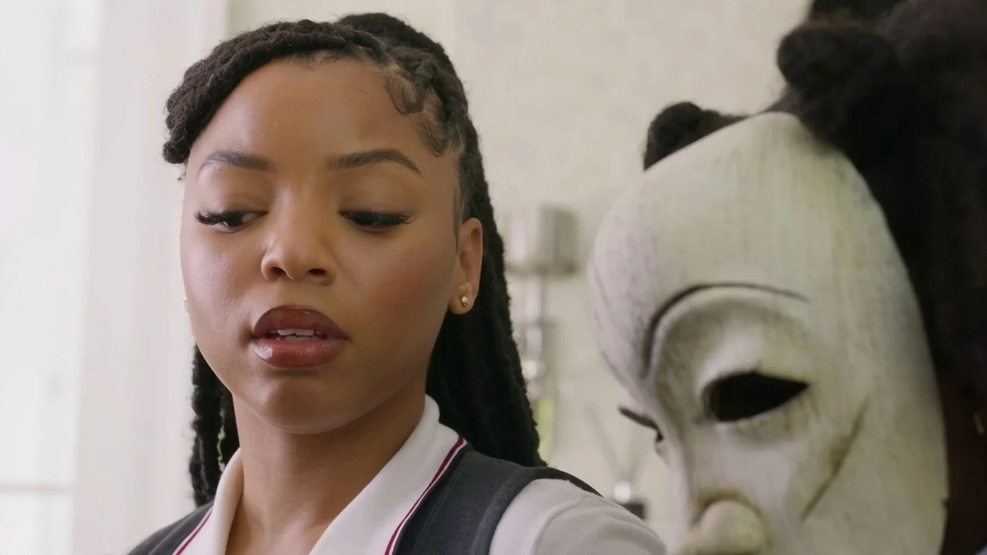 A figure in a white mask looms close beside Laila in Strung