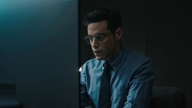 Rami Malek, as Charlie Heller, works at a computer in a dim office, his face lit by the screen, in The Amateur