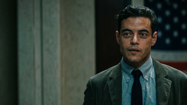 Rami Malek's character stands in a jacket and tie before a large American flag in a government office