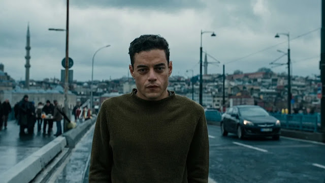 Rami Malek's character walks across a crowded bridge in Istanbul under a grey sky, mosques in the distance