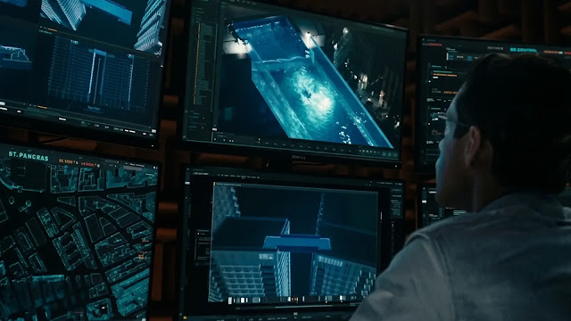 Rami Malek's character sits before a wall of glowing blue monitors showing maps and surveillance feeds