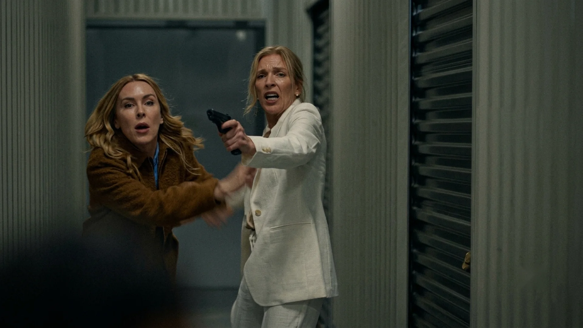 Clare points a gun at someone off-frame as Amy grabs at her arm, a desperate standoff in the storage corridor, in The Body in the Locker