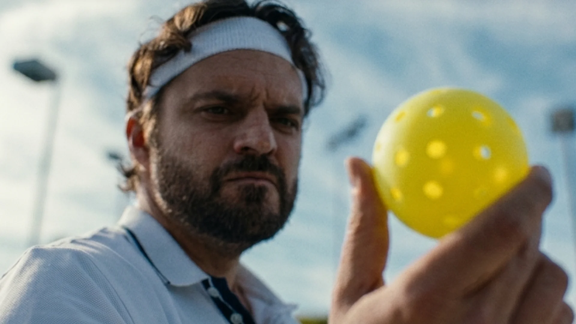 Jake Johnson, in a headband, holds a yellow pickleball up close and studies it with a skeptical look