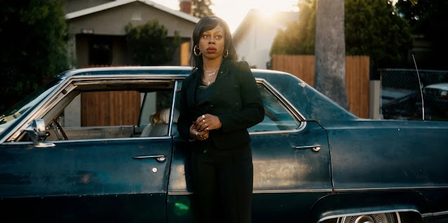 Wanda Bell (Gail Bean) standing beside a blue sedan on a South Los Angeles street in The Drop, late afternoon sun behind her