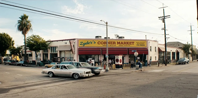 A corner market on a wide South Los Angeles street in The Drop, early 1990s cars parked along the curb and a palm tree above the roofline