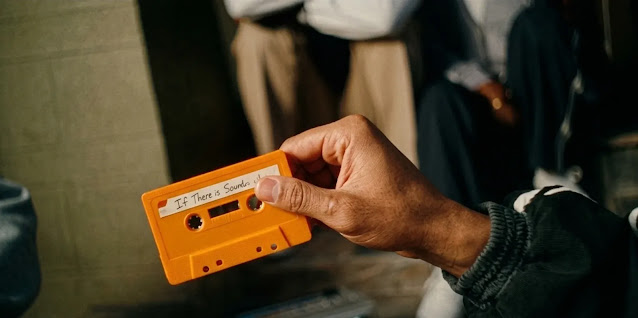A hand holding an orange cassette tape with a handwritten label in The Drop