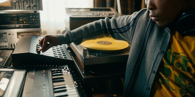 A young producer working a drum machine beside a turntable in The Drop, headphones around his neck and a record spinning