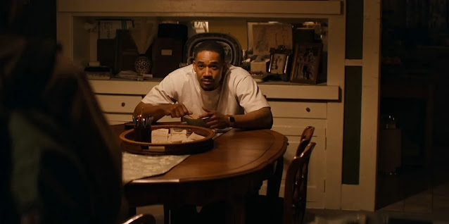 A man seated alone at a round dining table in The Drop, a plate in front of him and shelves of family things behind