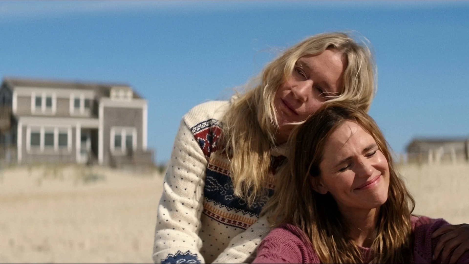 Two of the friends leaning into each other on a Nantucket beach, smiling, a beach house behind them