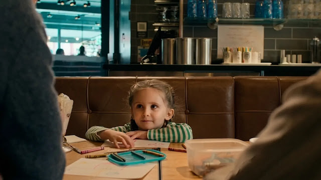 Ella (Grace Delaney) sits at a restaurant booth with crayons on the table in the We Live in Time trailer